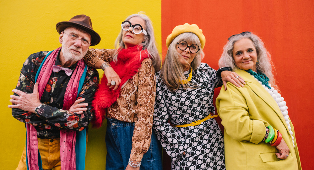 Active elderly people looking at the camera while standing together against a colourful wall. Group of four senior citizens feeling confident and youthful in colourful clothing
