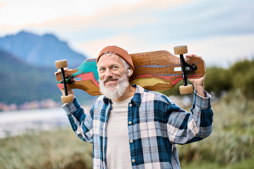 Active cool happy bearded old hipster man standing in nature park holding skateboard. Mature traveler skater enjoying freedom spirit and extreme sports hobby on mountains background. Portrait