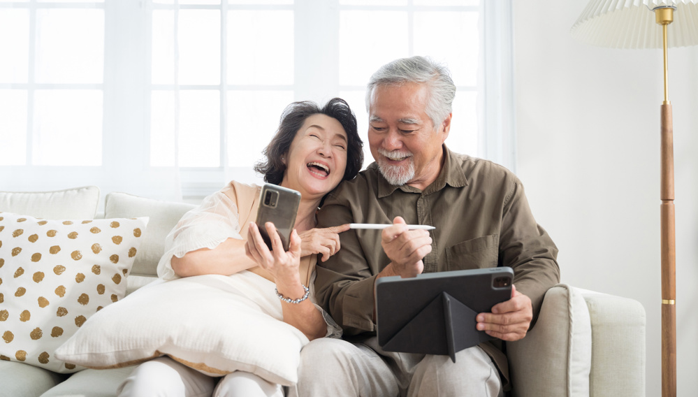 Asian senior couple in living room at home. Wife browsing online on smartphone showing something to her husband while husband is also using a tablet