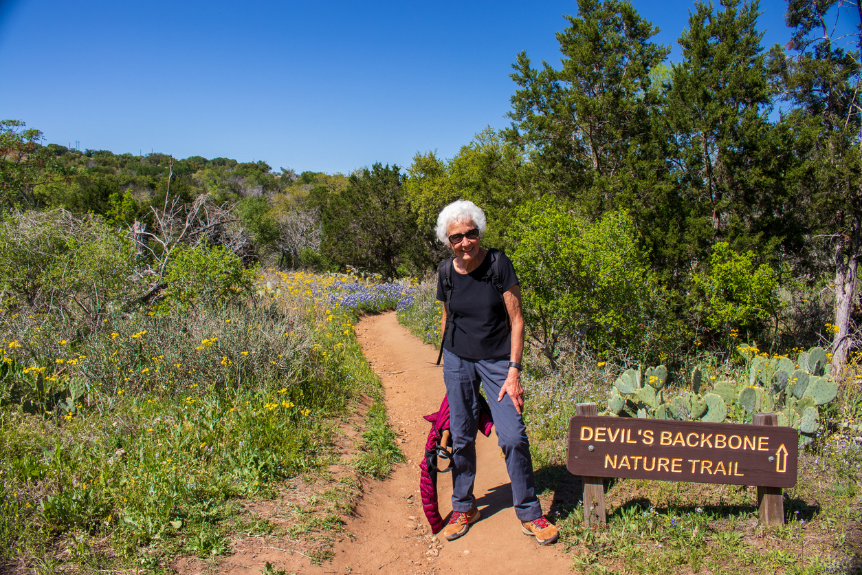 a senior woman posing in front of a trail sign with Texas Bluebonnets and wildflowers growing along a red dirt footpath part of the Texa Hill Country